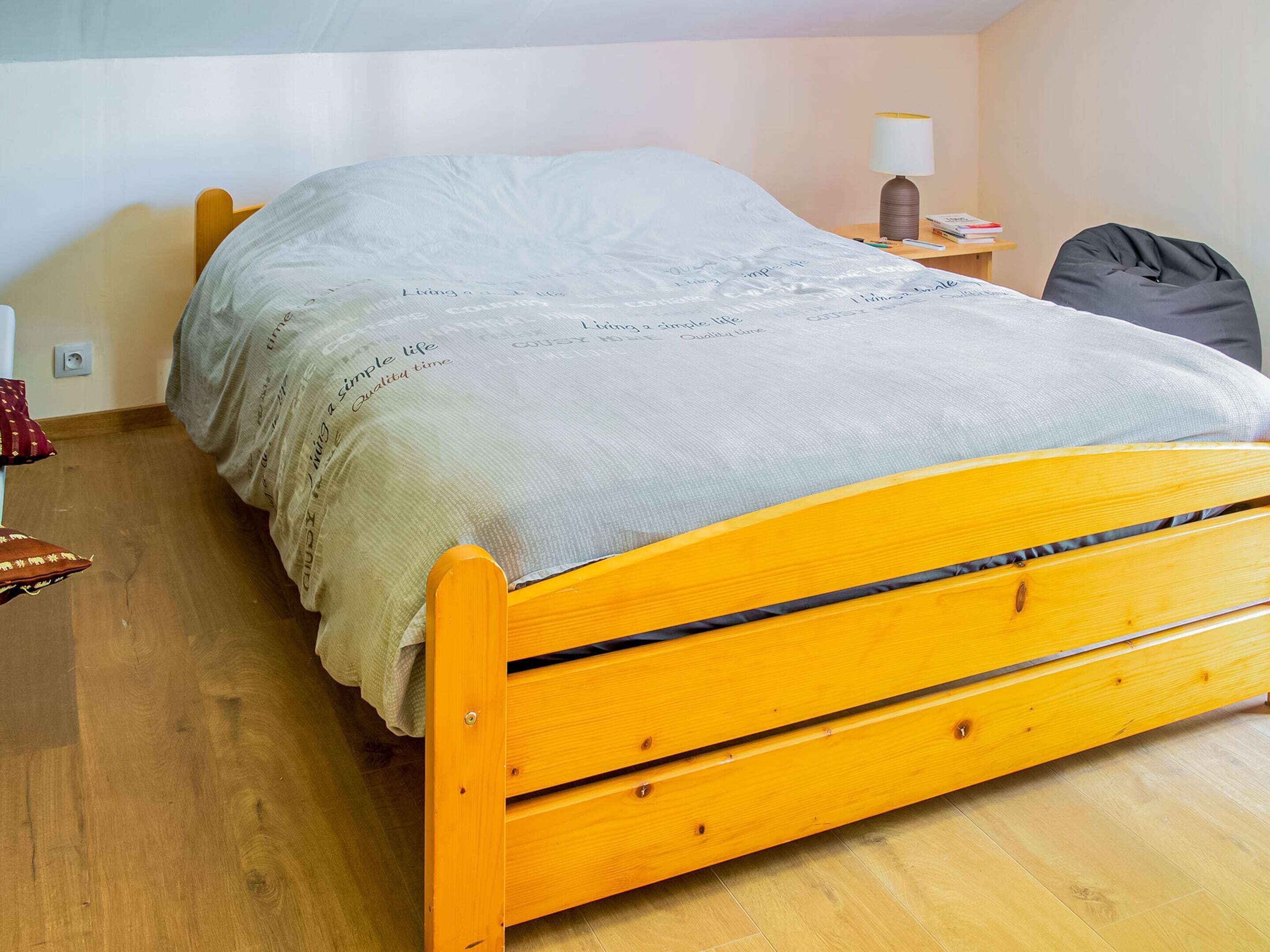 Un lit simple avec une tête de lit en bois, dans une pièce dotée d’un plancher en bois et d’une table de chevet sur laquelle se trouvent une lampe et des livres.