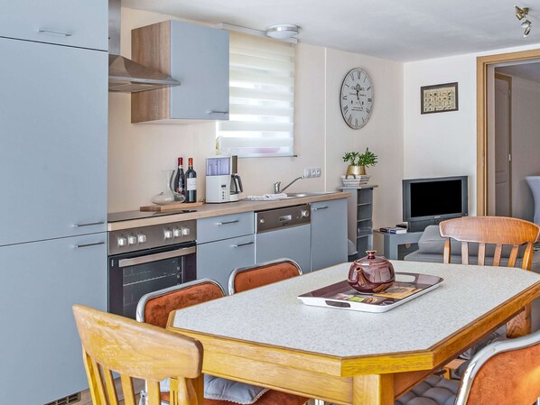A compact kitchen with modern appliances, a dining table with chairs, a clock, and a television. - Captivating Cottage in Vielsalm near Forest (Vielsalm)