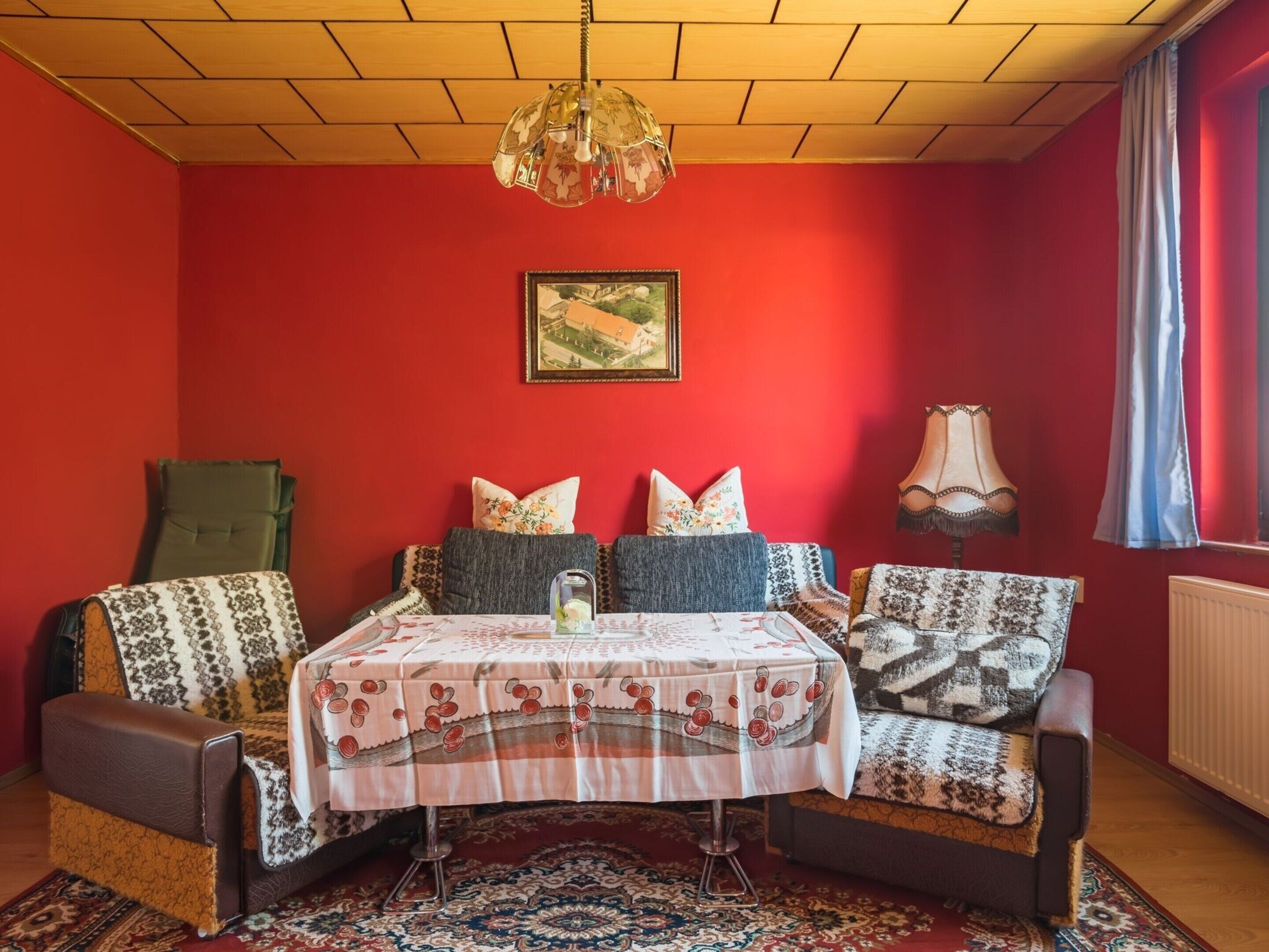 A cozy room with a red wall, a patterned rug, a table with a floral tablecloth, and a hanging lamp.