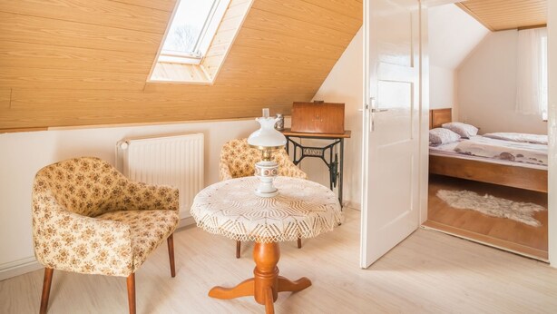 A cozy attic bedroom with a sloped ceiling, a skylight, a wooden table with a lace tablecloth, a vintage sewing machine, and a bed with a white blanket. - Countryside Stay in Meisdorf (Falkenstein)