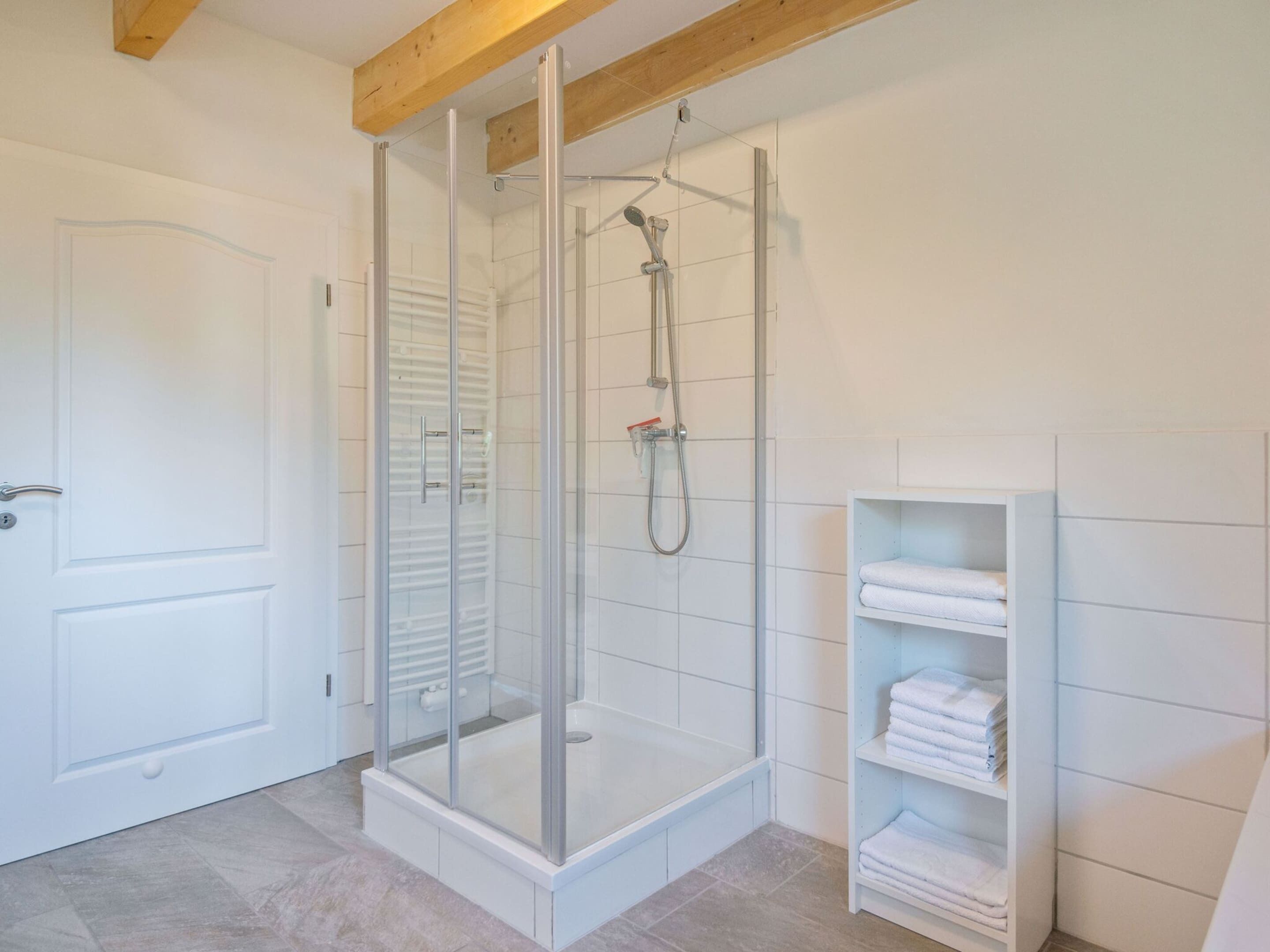 A modern bathroom with a glass-enclosed shower, white tiled walls, and a built-in shelf with folded towels.