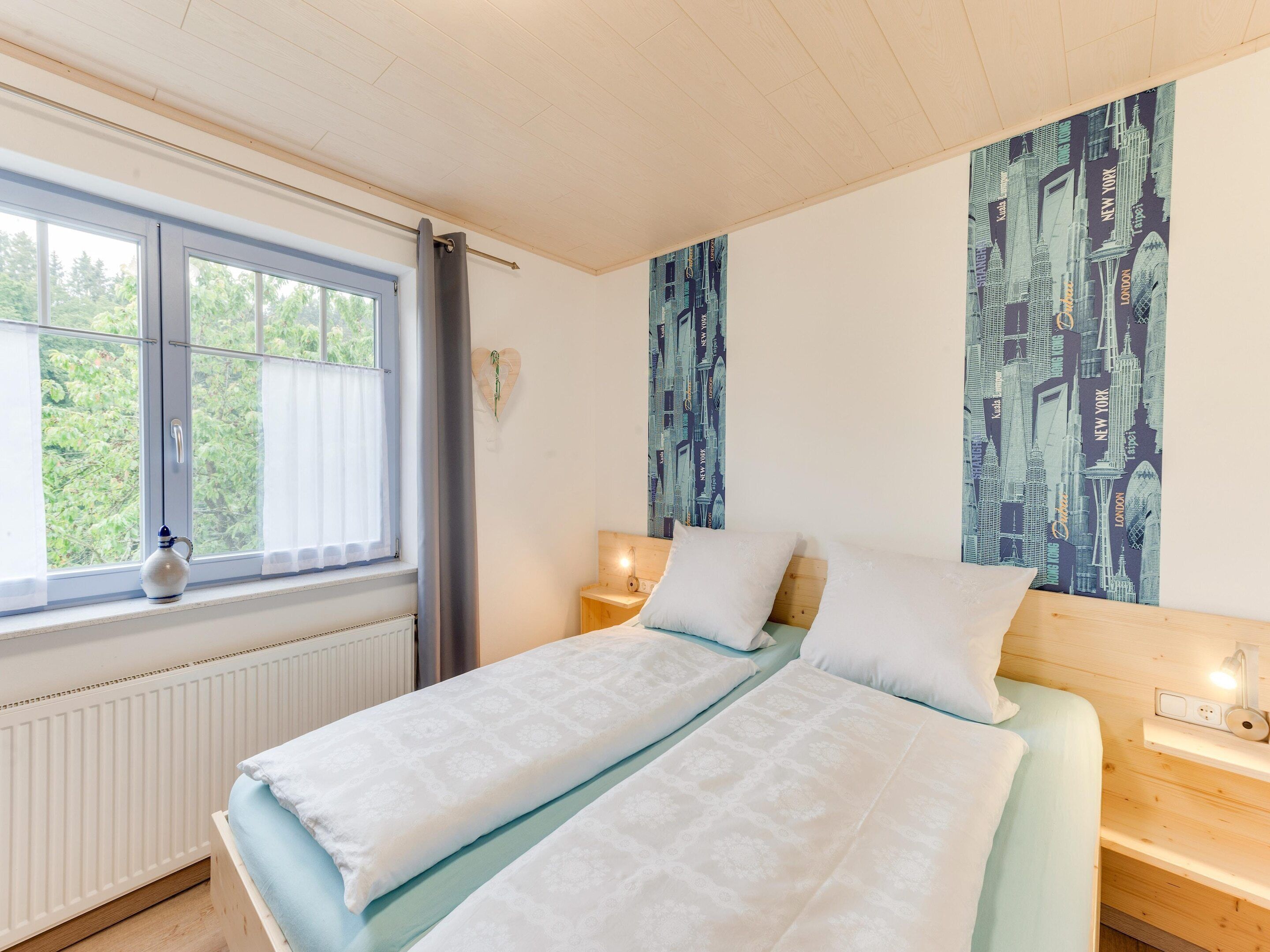 A bedroom with a bed, a window with a view of greenery, a wall with a cityscape print, and a bedside table with a lamp.