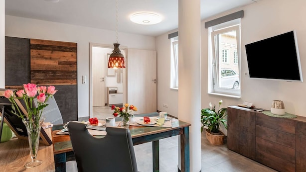 Un comedor moderno con una mesa de madera, sillas y un jarrón de flores. Se ve un televisor montado en la pared y una ventana con vistas a la calle.