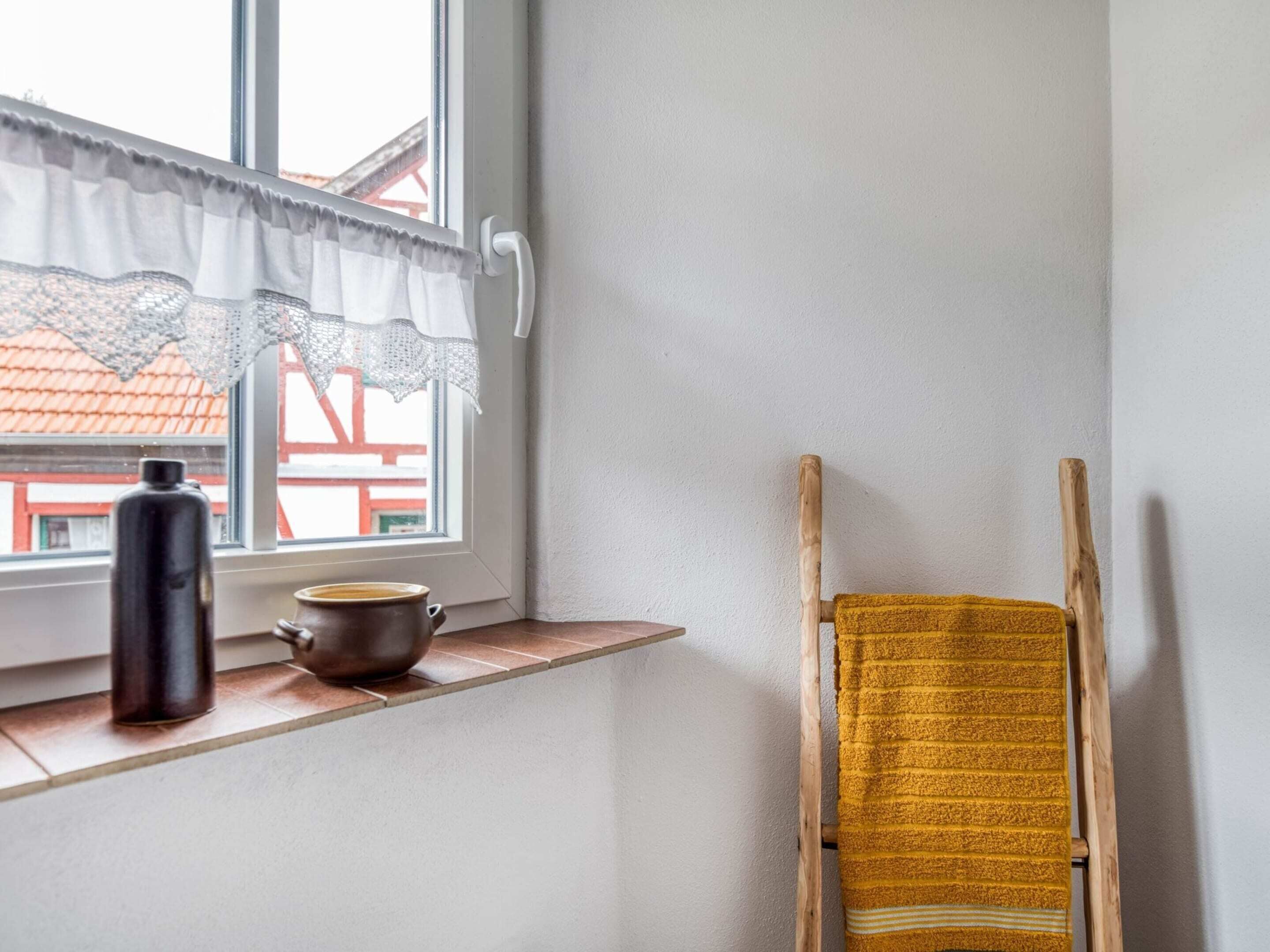 Een raam met een witte gordijn, een zwarte waterfles en een bruine pot op een plank. Een houten stoel staat tegen de muur.