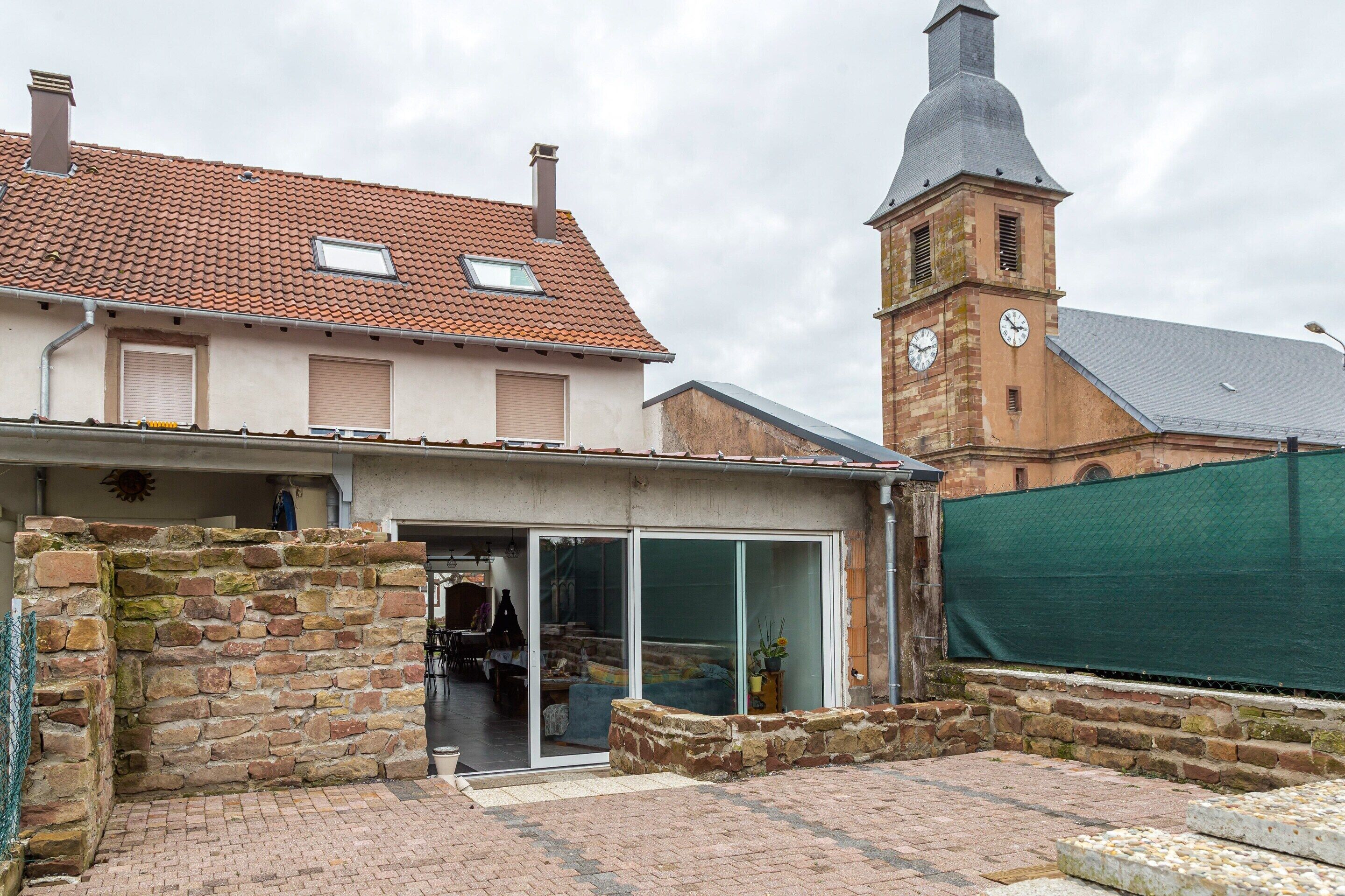 Un edificio tradicional con fachada de piedra, una torre de campanas y una extensión moderna con puertas de vidrio.