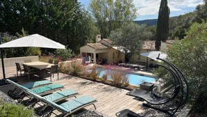 Una terraza de madera con sillones, piscina y un pequeño edificio con techo de tejas.