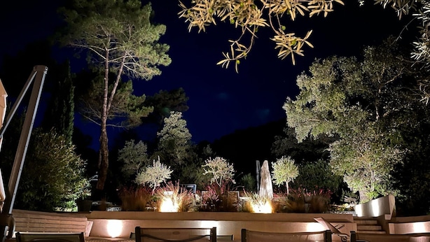 Un área de comedor al aire libre con sillas y mesas, rodeada de árboles y un elemento decorativo de iluminación.