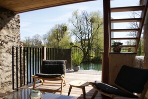 Une terrasse en bois avec deux fauteuils en osier, une petite table et une vue sur un étang et des arbres.