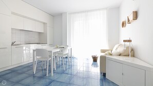 Een moderne keuken met witte kasten, een eetgedeelte met een witte tafel en stoelen, en een zithoek met een bank en een lamp.