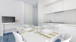 Een moderne, witte eetruimte met een flatscreen televisie aan de muur, een keuken met witte kasten, en een eettafel gedekt met borden, glazen en bestek.