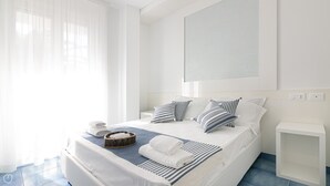 Una habitación de hotel moderna con una cama grande, ropa de cama blanca, almohadas a rayas, una mesita de noche pequeña y un ventanal con cortinas translúcidas.