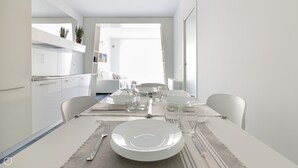 Un comedor moderno con una mesa preparada para cuatro personas, sillas blancas y una cocina visible al fondo.