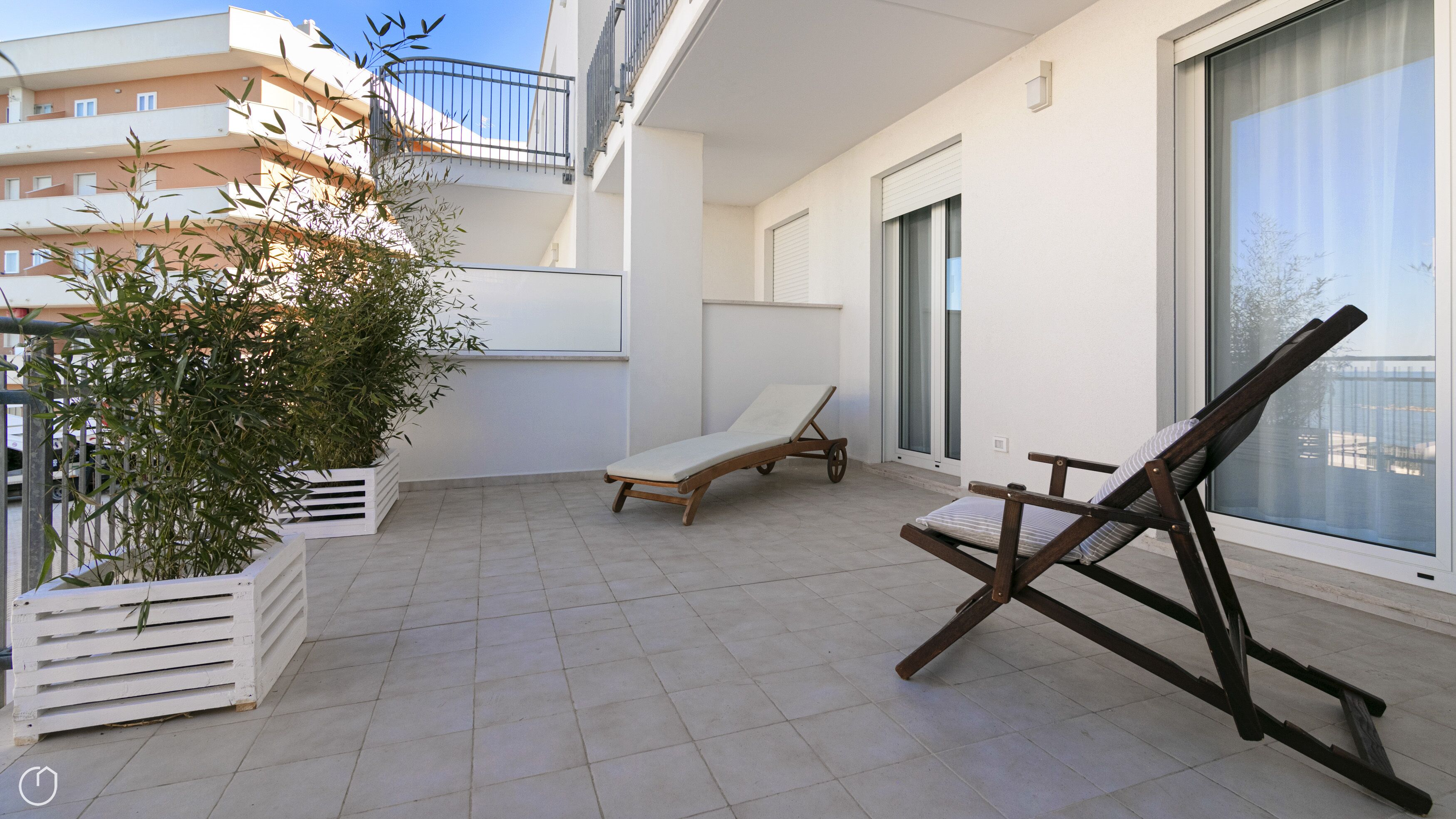 A balcony with a white tiled floor, a wooden lounge chair, a sun lounger, and a potted plant.