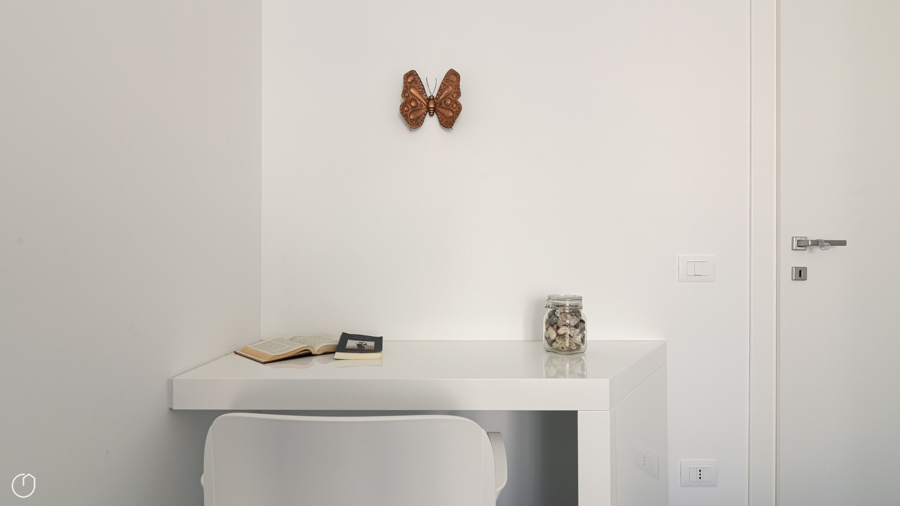 A minimalist room with a white desk, a book, a decorative butterfly, and a jar of seashells.