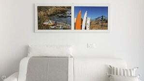 Una cama con ropa de cama blanca y un plaid gris, una pared blanca, y dos cuadros que muestran un paisaje fluvial rocoso y una playa con tablas de surf.