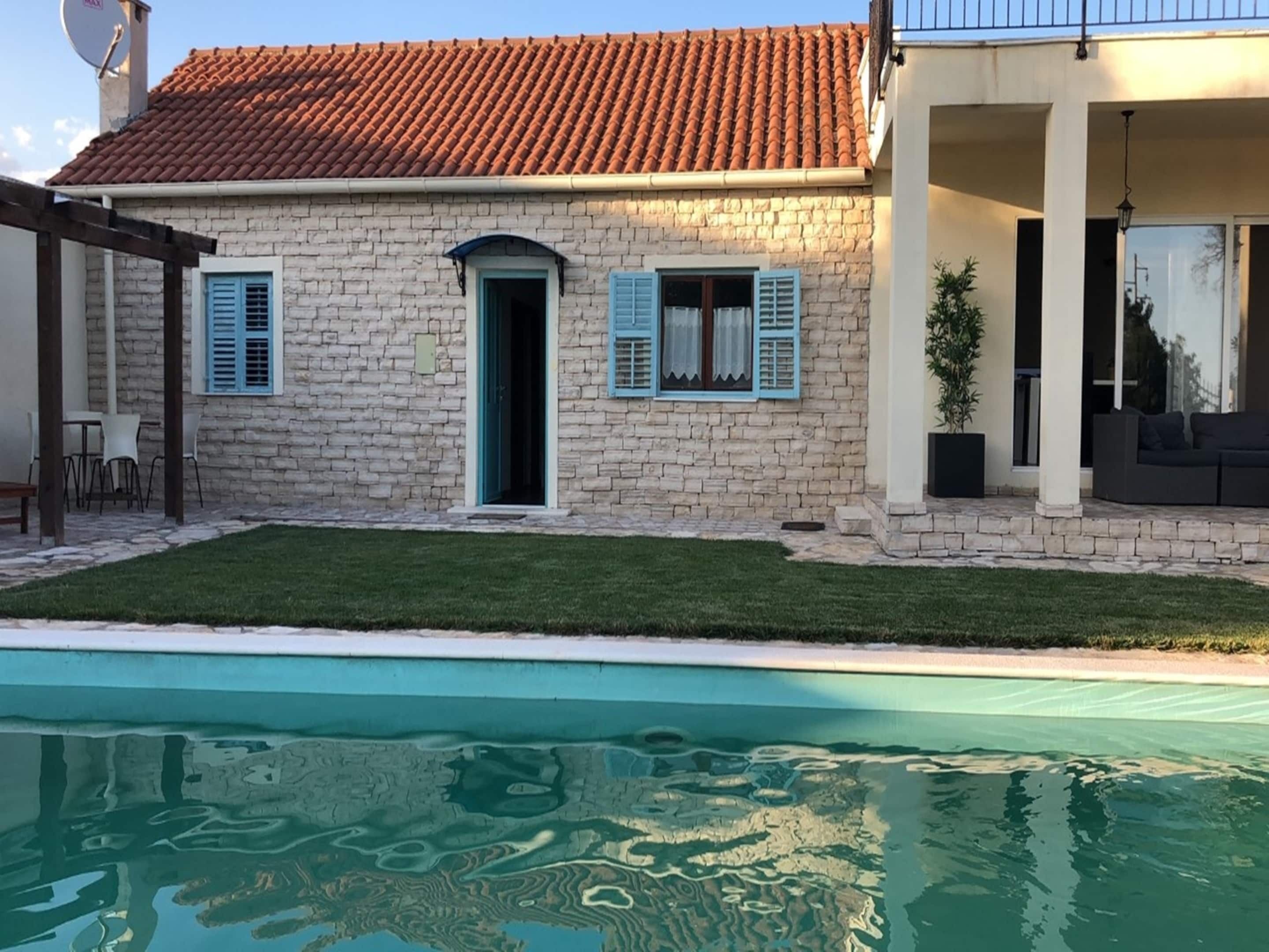 A stone house with a swimming pool, a door, and two windows with shutters.