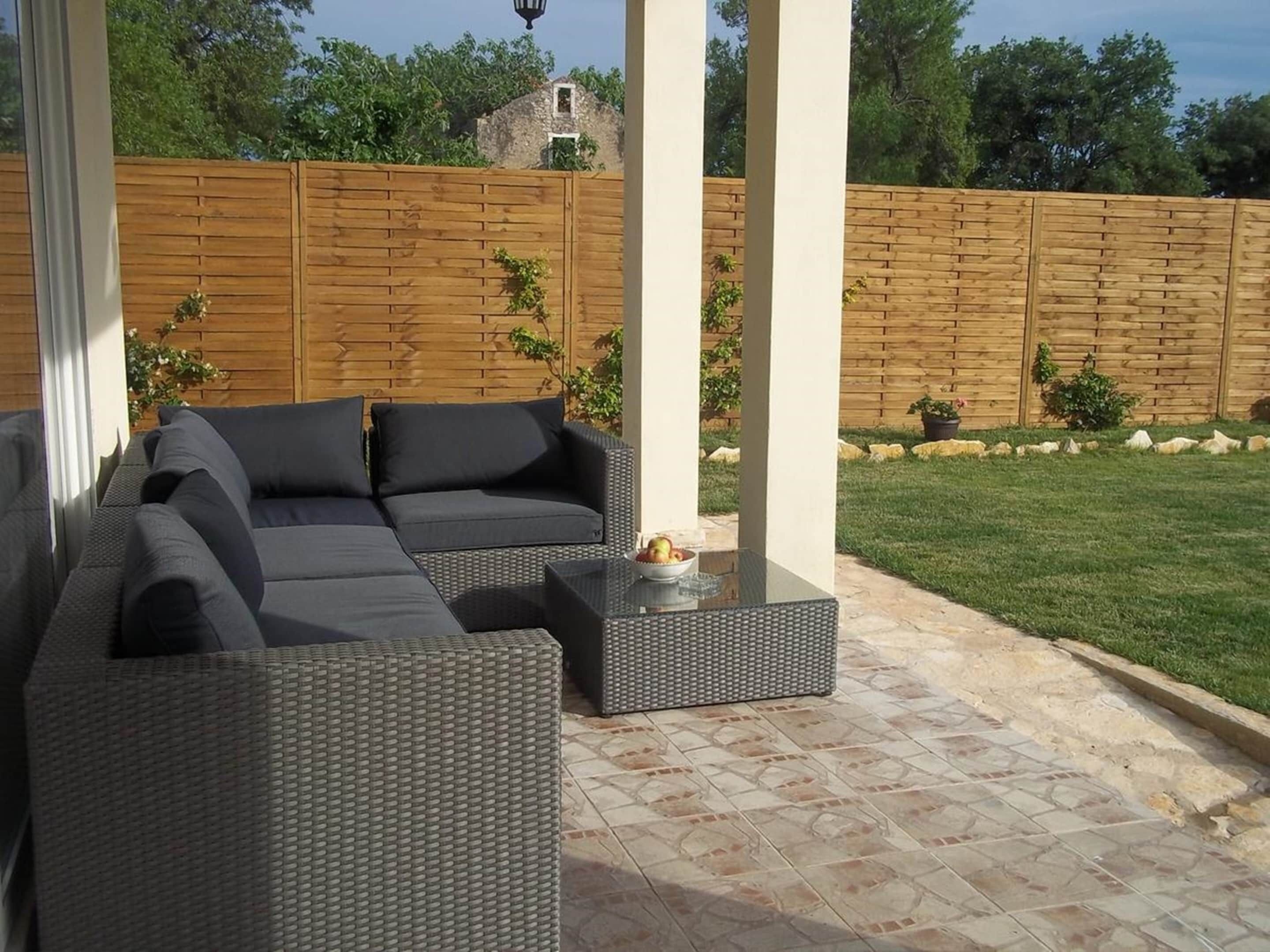 A patio area with a sectional sofa and a small table, surrounded by a wooden fence and a grassy lawn.