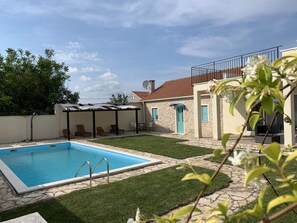Una piscina con un pérgola, una casa de piedra con puerta azul y un jardín con césped y plantas.