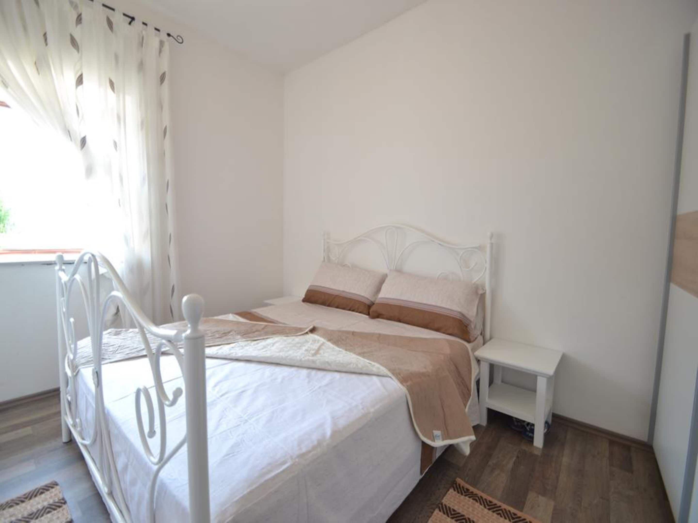 A bedroom with a white metal bed frame, a white mattress, and a brown and white striped blanket. There is a white bedside table with a lamp and a small rug on the floor.