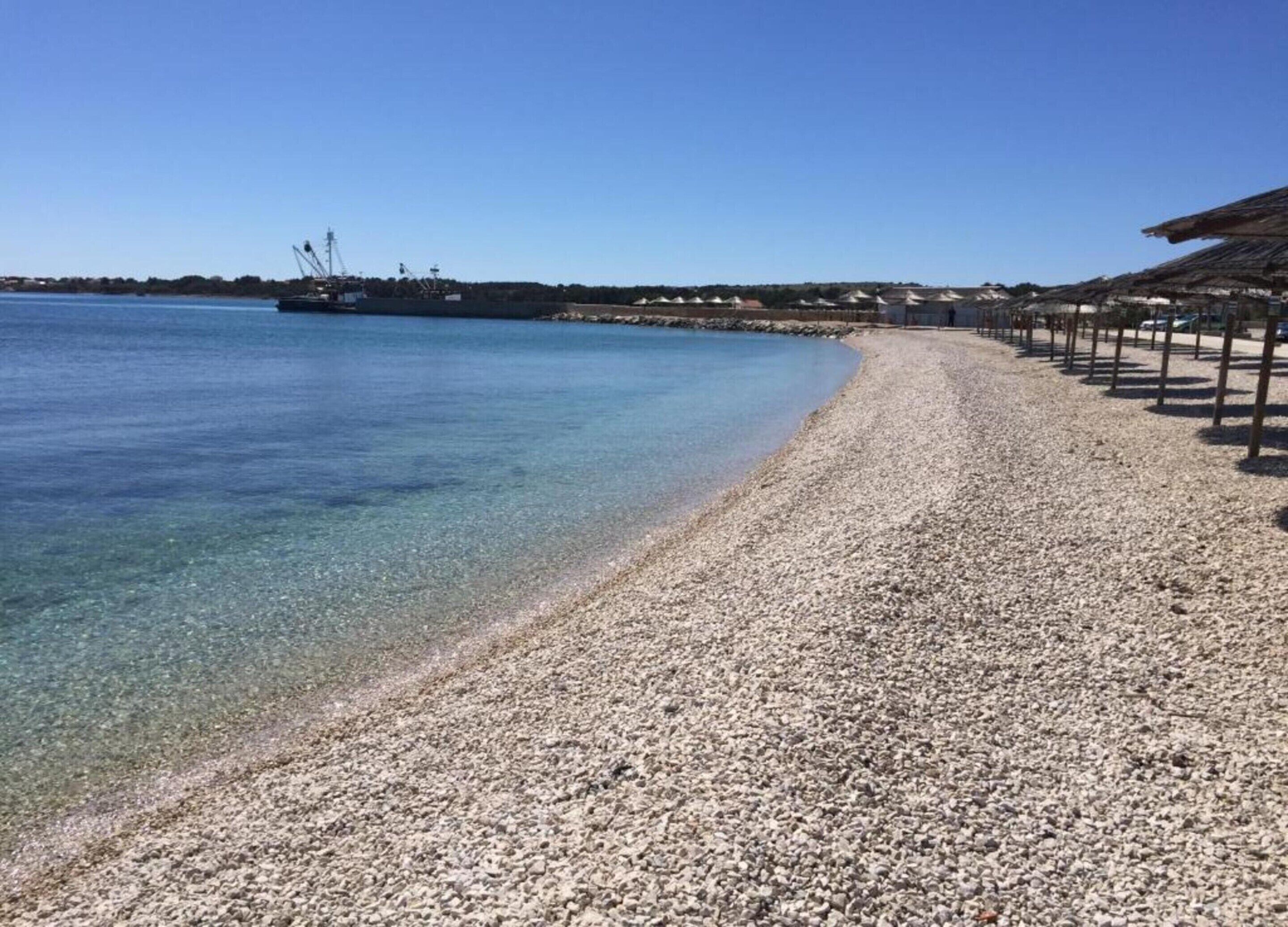 Une plage de galets bordée d’une étendue d’eau claire, un grand navire visible au loin et une rangée de bâtiments sur la droite.