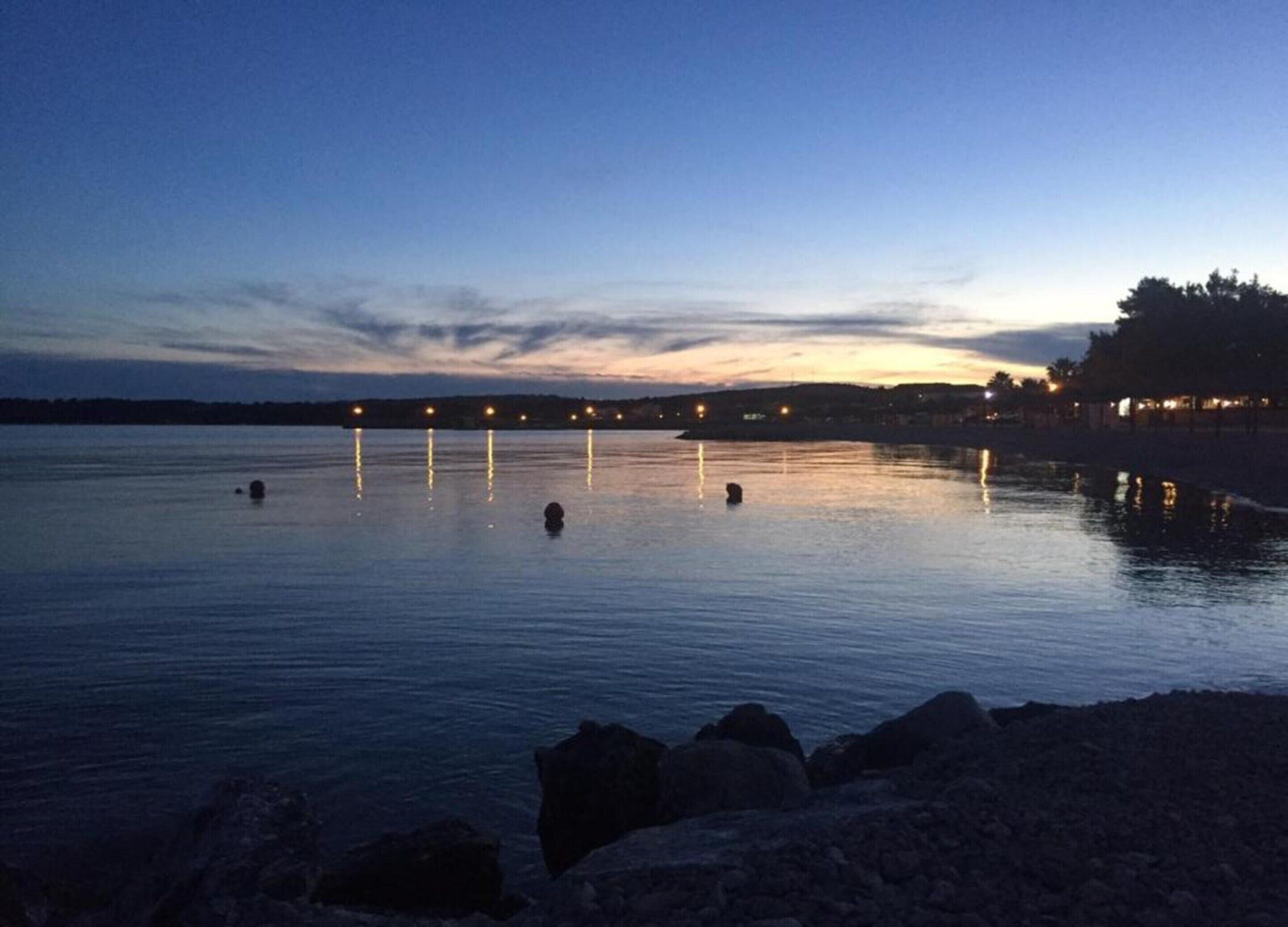 A calm waterfront at dusk with a row of evenly spaced lights reflecting on the water.