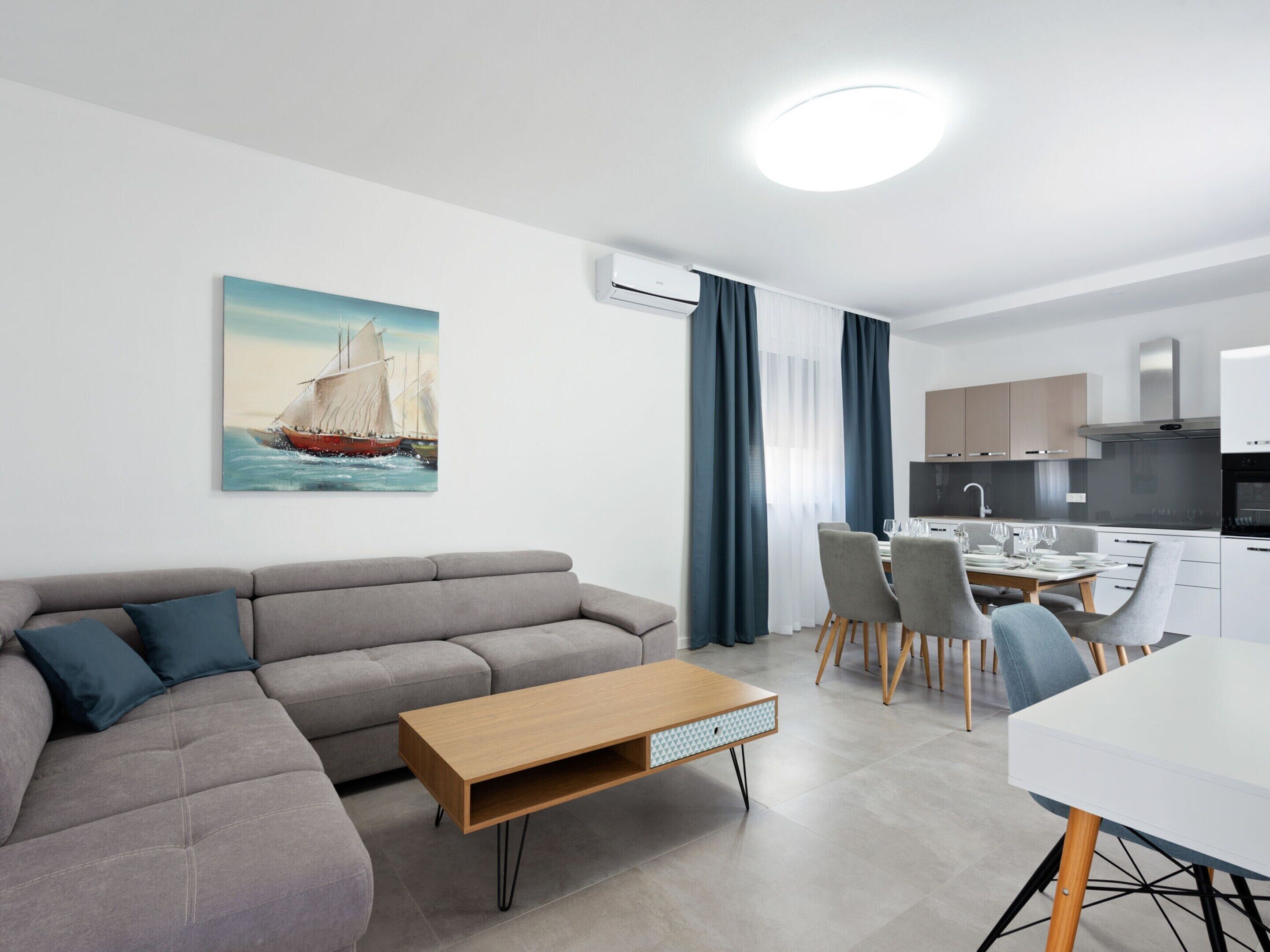 Een moderne woonkamer met een grijze bank, een houten salontafel, en een eethoek met een witte tafel en stoelen.