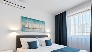 A hotel room with a bed, a painting of sailboats, a window with curtains, and an air conditioning unit. - Seaside Comfort on Vir Island (Vir)
