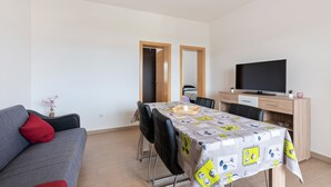 Un salon moderne avec un canapé gris, une table à manger recouverte d’une nappe à motifs, un téléviseur à écran plat et un bureau en bois.