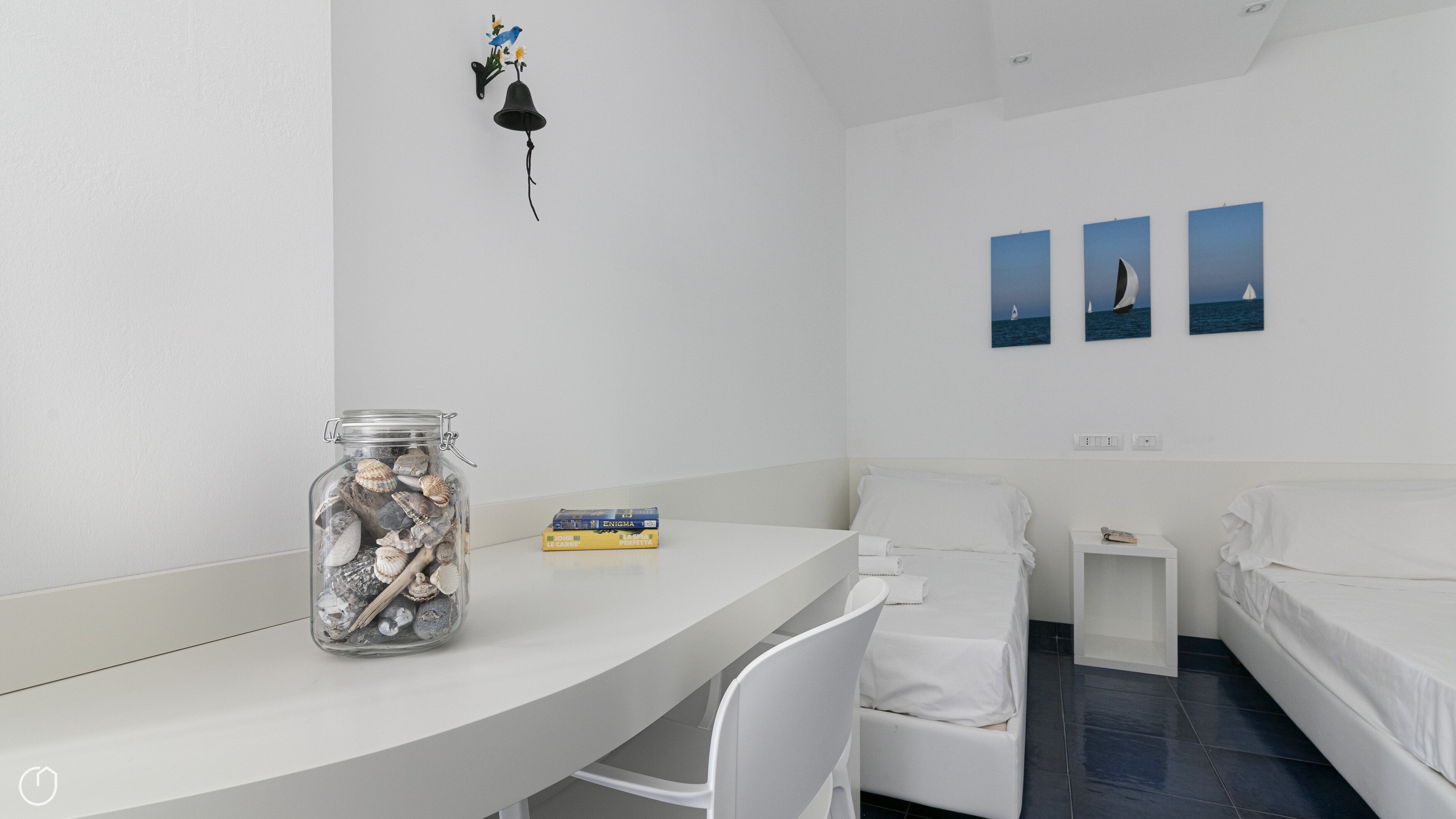 Una habitación con un escritorio blanco, un tarro de vidrio con caracoles, una pequeña estantería con un libro, dos camas blancas y tres cuadros en la pared.