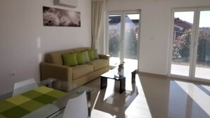Un salon moderne avec un canapé beige, une table à manger en verre et une grande fenêtre donnant sur un quartier résidentiel.