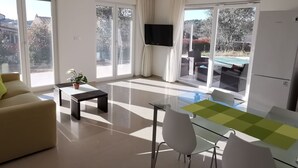 Un salon moderne comprenant un canapé, une table basse avec une plante, une table à manger en verre avec des chaises, un téléviseur à écran plat et de grandes portes-fenêtres coulissantes donnant sur un balcon avec vue sur la piscine.