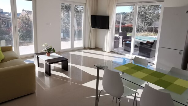 Un salon moderne comprenant un canapé, une table basse avec une plante, une table à manger en verre avec des chaises, un téléviseur à écran plat et de grandes portes-fenêtres coulissantes donnant sur un balcon avec vue sur la piscine.