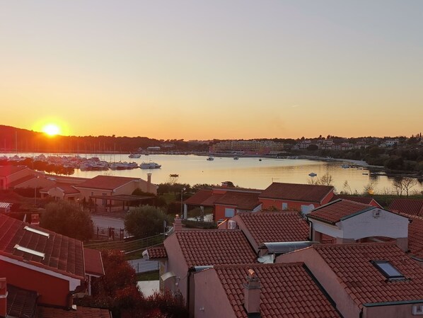 A coastal town at sunset with boats docked along the harbor, residential buildings with red-tiled roofs, and a bridge in the background.