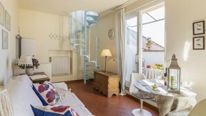 Ein gemütliches Wohnzimmer mit einer weißen Couch, gemusterten Kissen, einem Holznachttisch, einer Lampe, einem kleinen Tisch mit Glasplatte und einer Wendeltreppe.