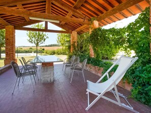 Une terrasse couverte, dotée d’une table et de chaises, entourée de verdure et offrant une vue sur la campagne.