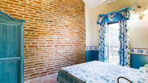 A room with a brick wall, a bed with a floral bedspread, a blue door, and a window with blue curtains. - Holiday Home in Castiglione Near the Lake (Castiglione del Lago)