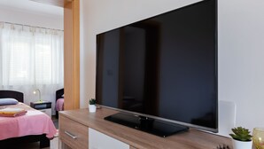 A modern bedroom with a large flat-screen TV mounted on a wooden dresser, a bed with a pink bedspread, a small bedside table with a lamp, and a potted plant. - Simplistic Apartment in Neviđane near Sea (Pasman)