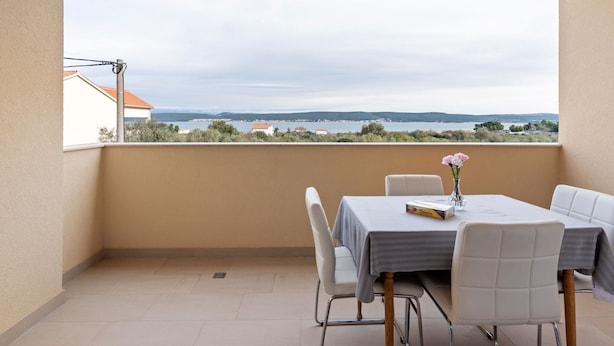 A balcony with a table and chairs, a vase with flowers, and a view of a body of water and distant landscape. - Simplistic Apartment in Neviđane near Sea (Pasman)