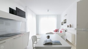 A modern, minimalist kitchen with white cabinetry and a built-in stove. There is a dining area with a white table and chairs, and a sofa with red cushions. - Rivazzurra Homes - 55 (Termoli)