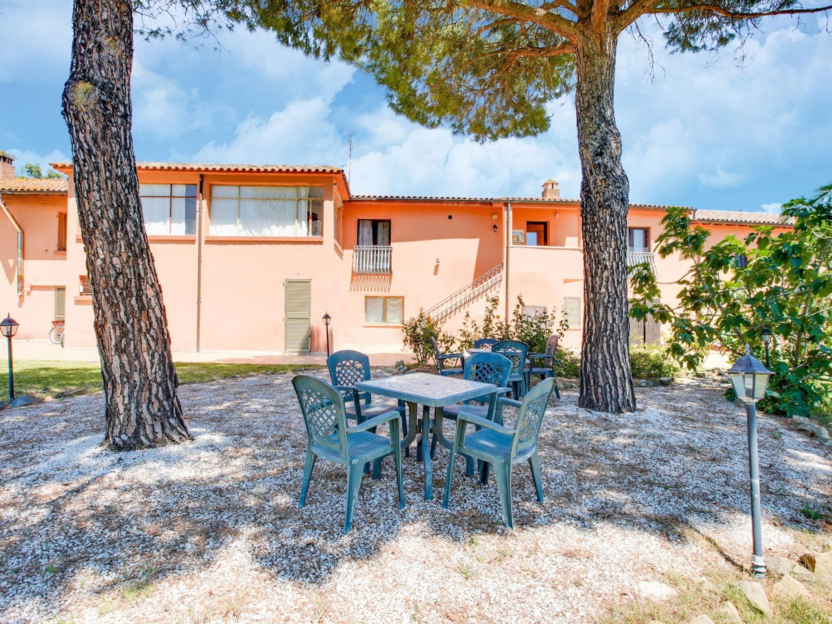 Un bâtiment à la façade rouge, avec un espace extérieur aménagé avec des chaises et une table, et deux grands arbres au premier plan.