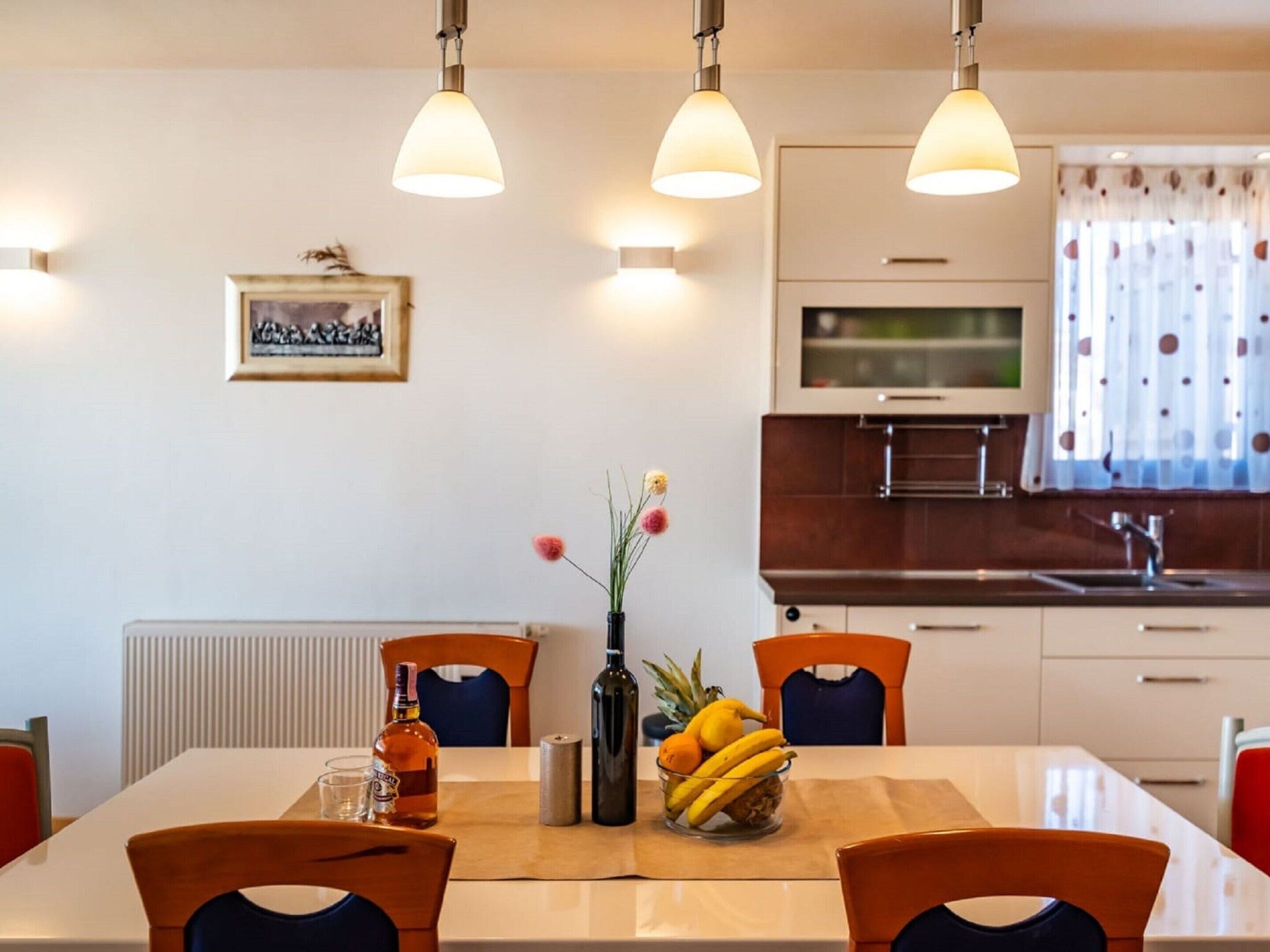 A modern kitchen with white cabinets, a built-in oven, and a stainless steel sink. There is a dining table with chairs and a vase with flowers.