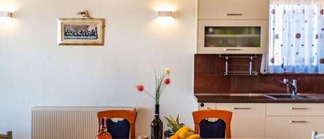 A modern kitchen with white cabinets, a built-in oven, and a stainless steel sink. There is a dining table with chairs and a vase with flowers.