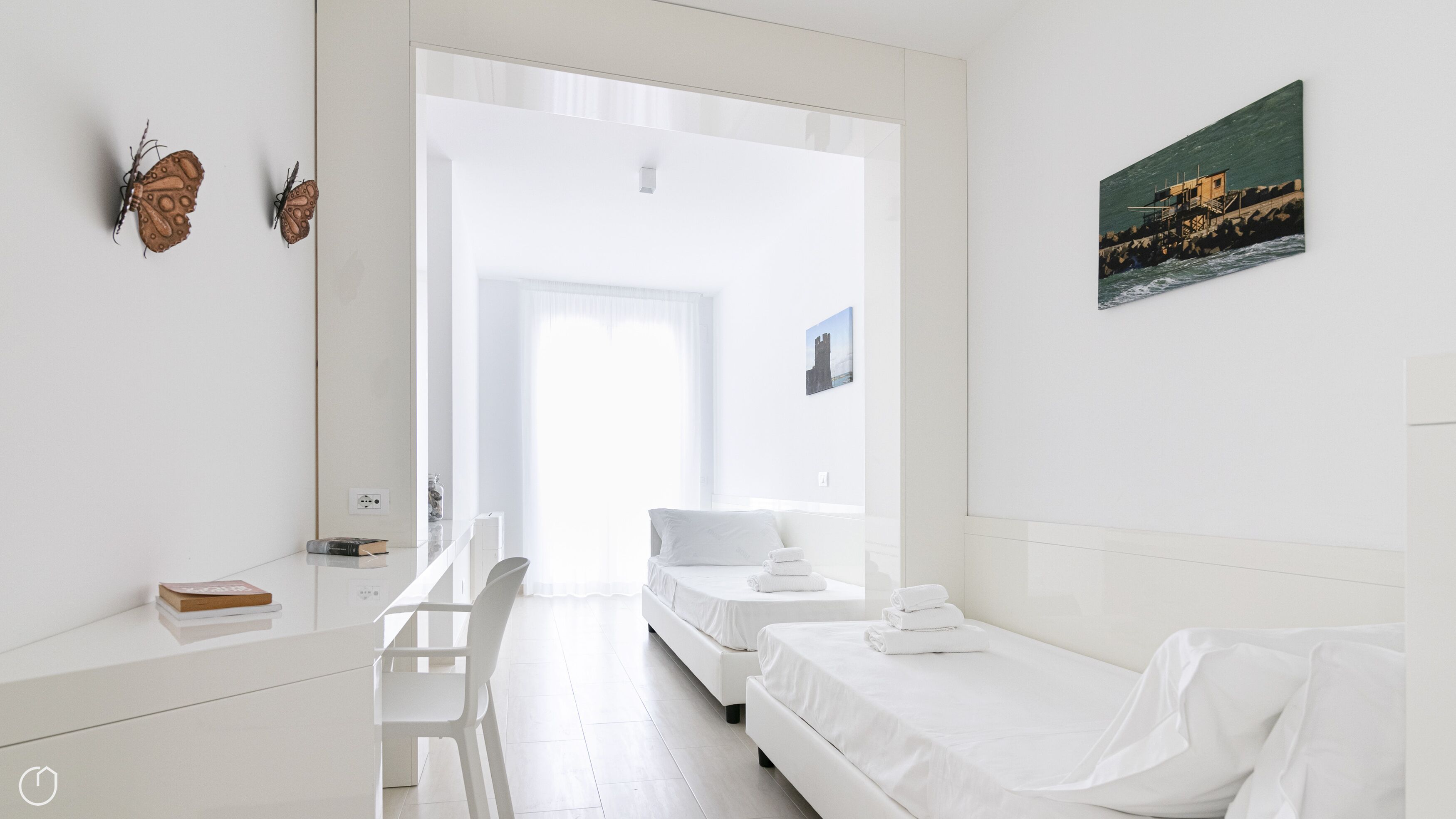 Ein helles, weißes Hotelzimmer mit einem Bett, einem Schreibtisch und einem Sessel. An der Wand hängt ein Gemälde und es gibt ein Fenster mit Vorhängen.