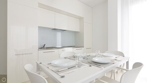 A modern, white dining area with a kitchen in the background, featuring a stainless steel sink and cabinets. - Rivazzurra Homes - 56 (Termoli)