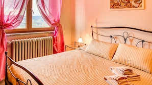 A bedroom with a bed, a radiator, a window with curtains, a bedside table with a lamp, and a painting on the wall.