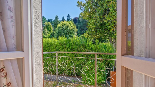 A balcony with ornate metal railings, a tiled floor, and a view of a lush garden with various trees and shrubs. - Belvilla by OYO Belle Epoque (Meina)