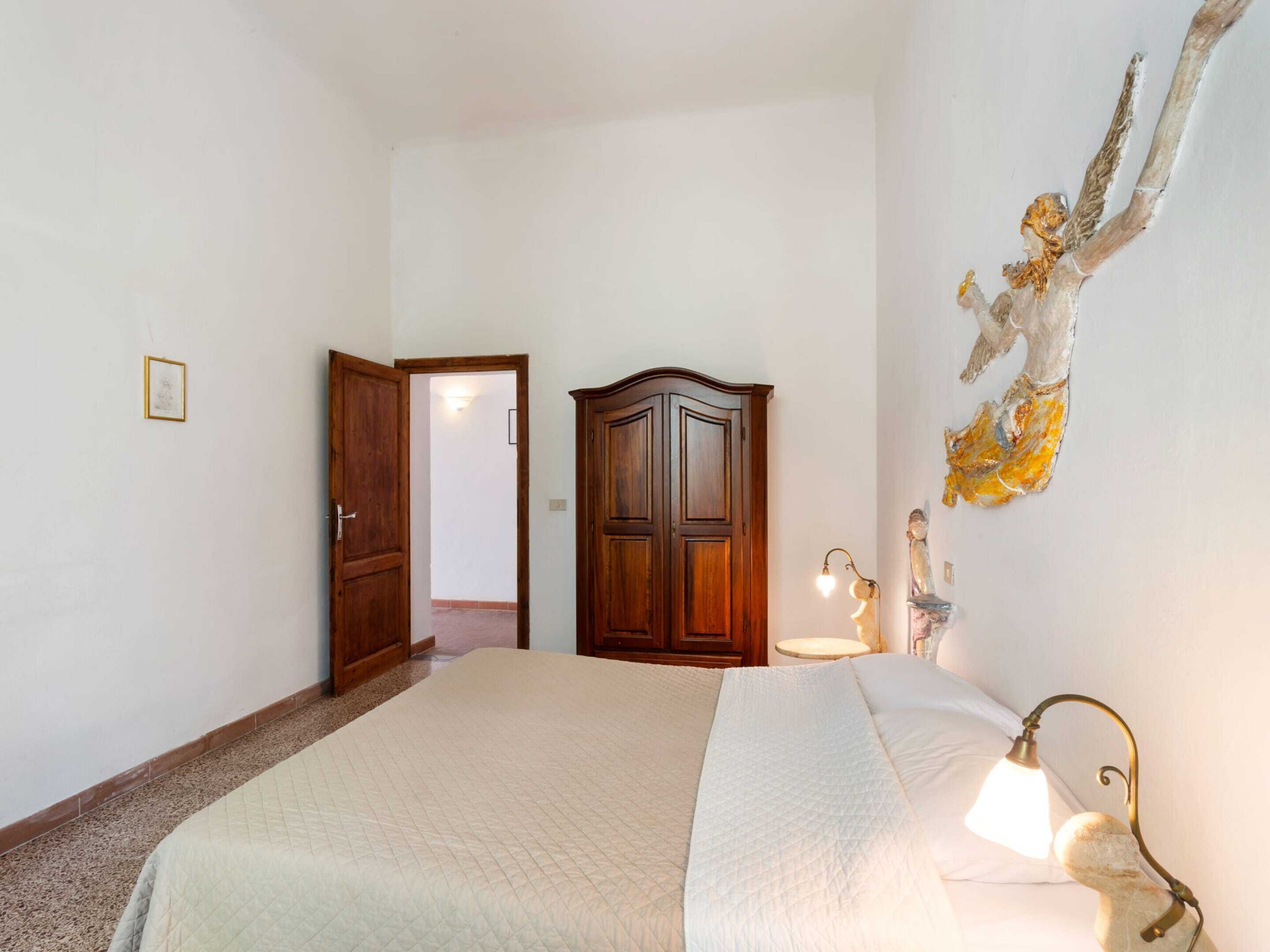 A bedroom with a large bed, a wooden wardrobe, a wall-mounted lamp, and a door leading to another room.