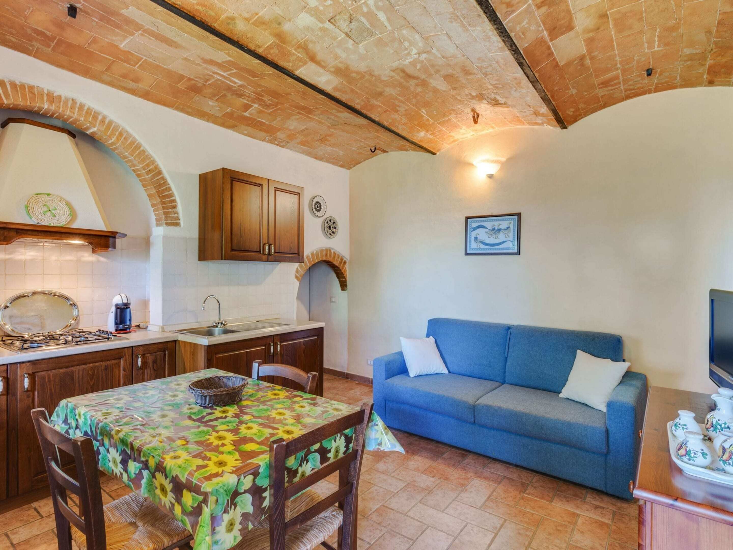 A cozy living area with a blue sofa, a dining table with a floral tablecloth, and a kitchen with wooden cabinets and a brick ceiling.