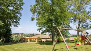 A grassy outdoor area with a wooden swing set, a covered seating area, and a view of distant buildings. - Holiday Home in Volterra With Pool (Volterra)