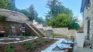 Ein Garten mit einer Steinmauer, einer Holzterrasse mit Kissen und einer Treppe zu einem überdachten Bereich.